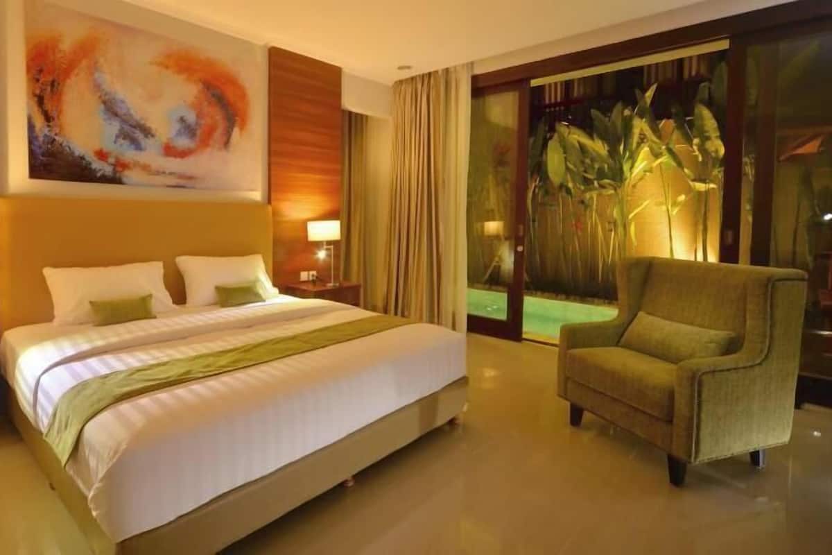 A hotel room with a large bed, two bedside tables with lamps, a large abstract painting, a chair, and a view of a tropical landscape through a glass door.