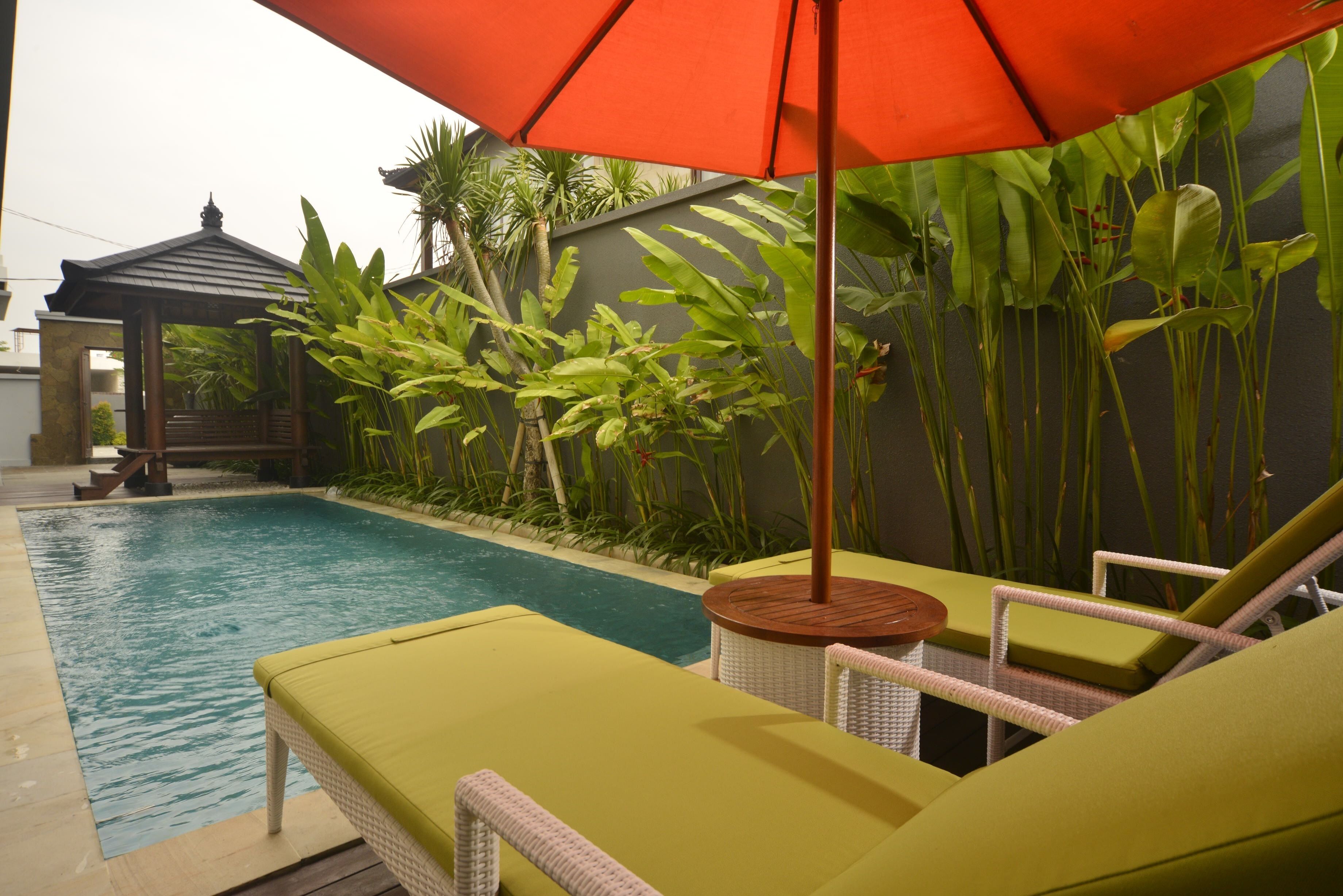 A poolside area with lounge chairs and an umbrella, surrounded by lush greenery.