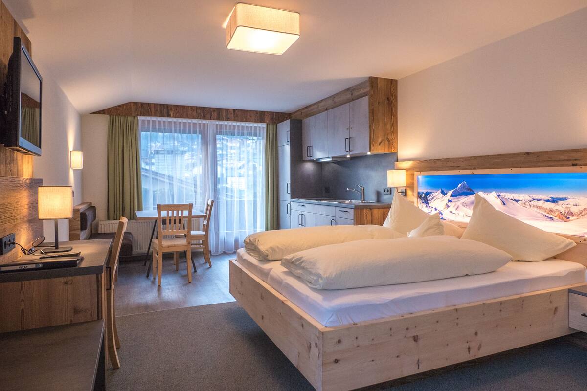 Ein Hotelzimmer mit einem großen Bett, einem Schreibtisch mit Stuhl, einer kleinen Küchenzeile und einem großen Fenster mit Bergblick.