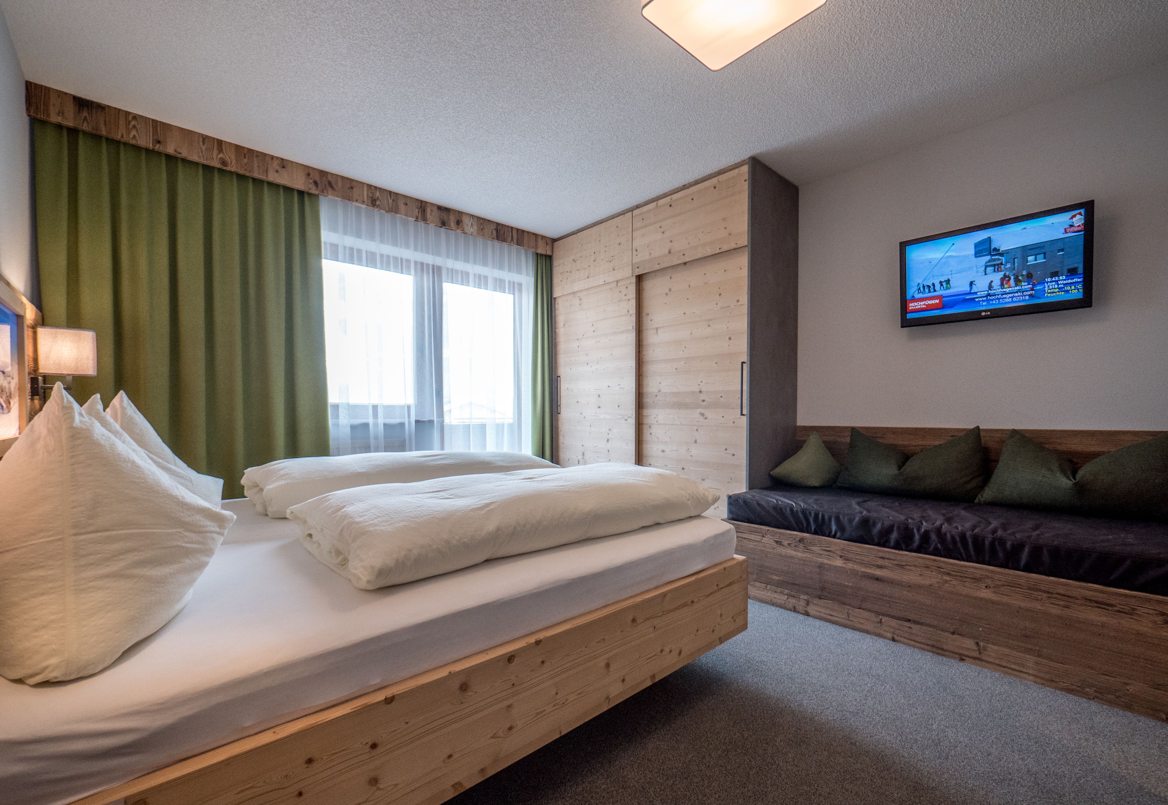 A modern hotel room with a wooden bed, a TV mounted on the wall, and a green curtain.