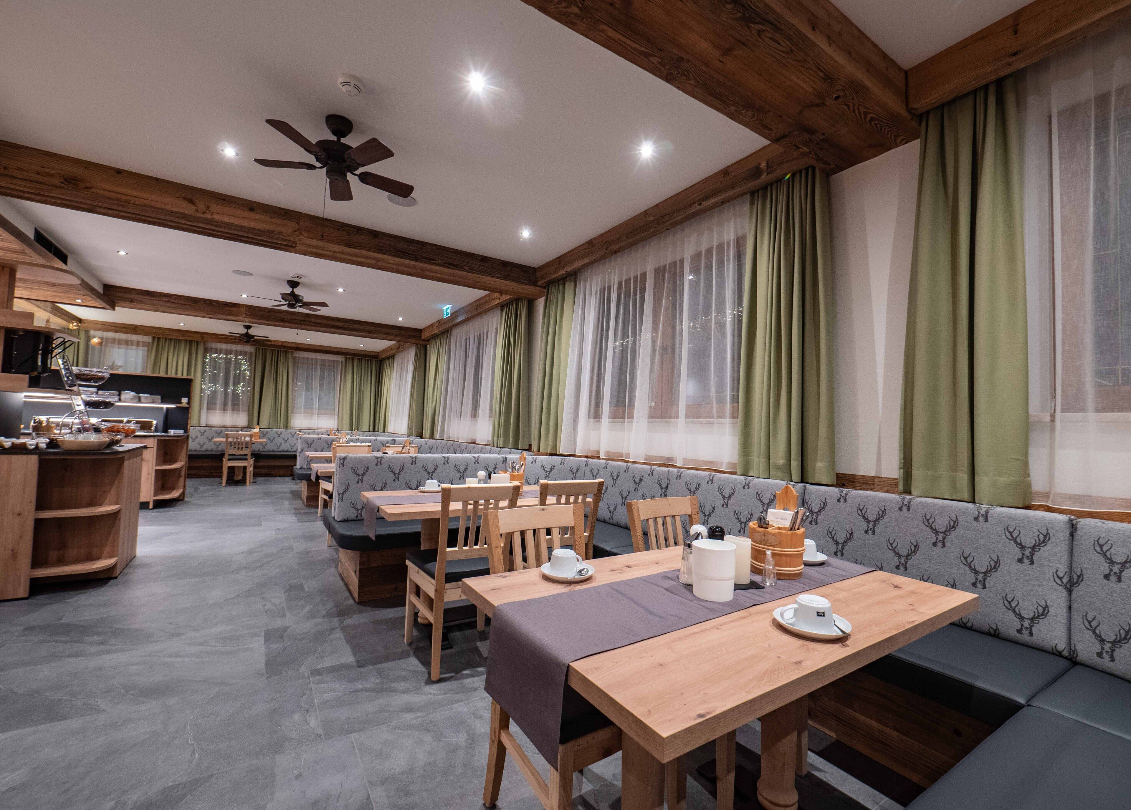 A restaurant with wooden tables and benches, a kitchen area, and large windows with green curtains.