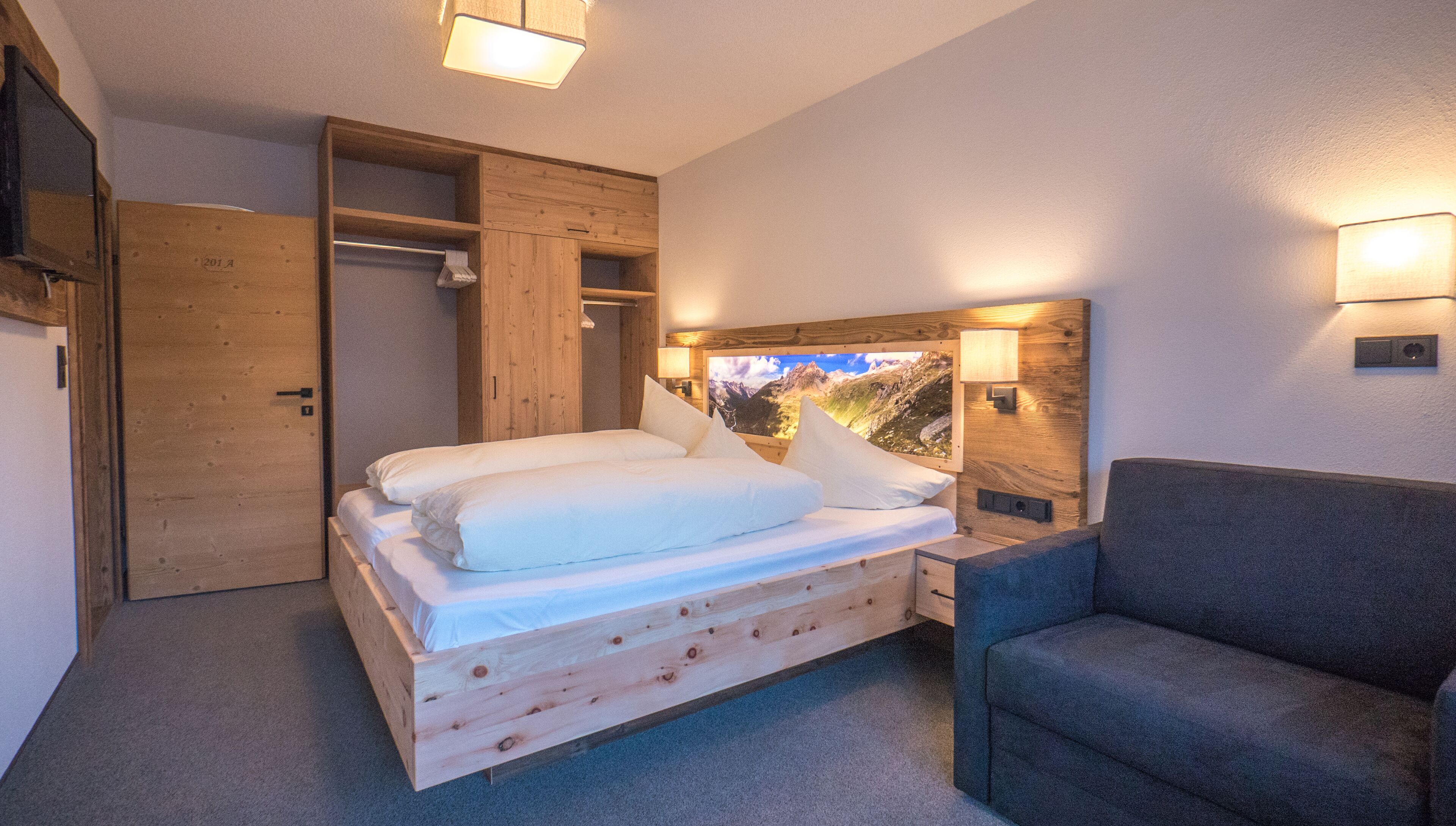 Ein hölzernes Bett mit Kopfteil, ein Nachttisch und ein grauer Sessel in einem Zimmer mit Holzwänden und Blick auf die Berge am Kopfteil.