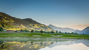 Een rustige golfbaan met een helder meer dat het landschap weerspiegelt, omringd door bergen en enkele gebouwen.