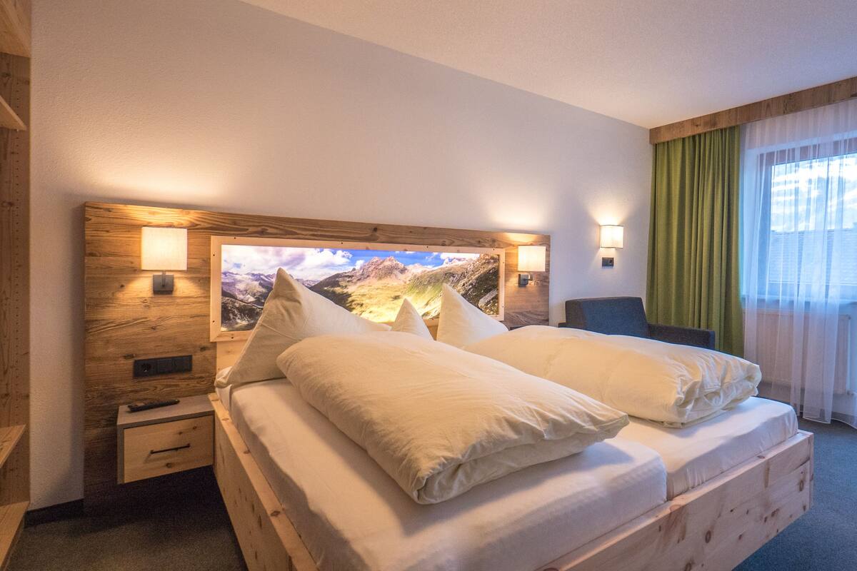 Ein Hotelzimmer mit einem hölzernen Kopfteil, einem Bett mit weißer Bettwäsche, einem Nachttisch, einer Lampe und einem Fenster mit durchsichtigen Vorhängen.