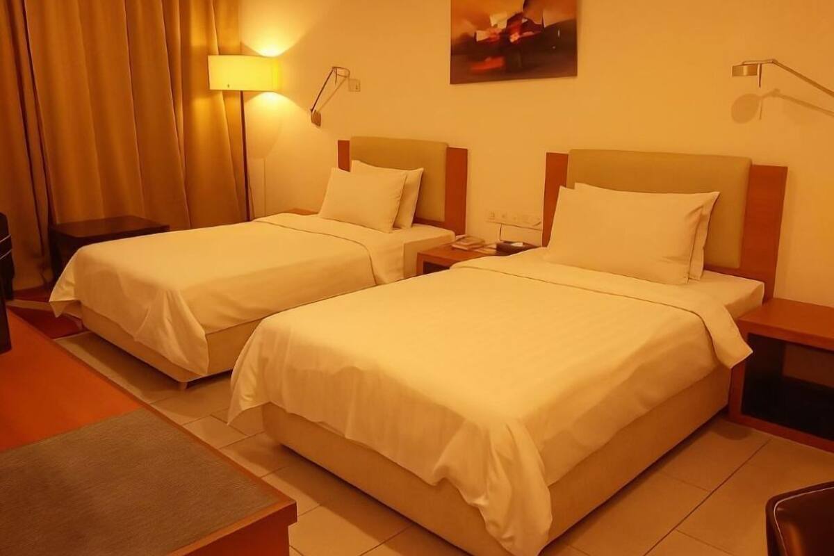A hotel room with two beds, a desk, a chair, a lamp, and a painting on the wall.