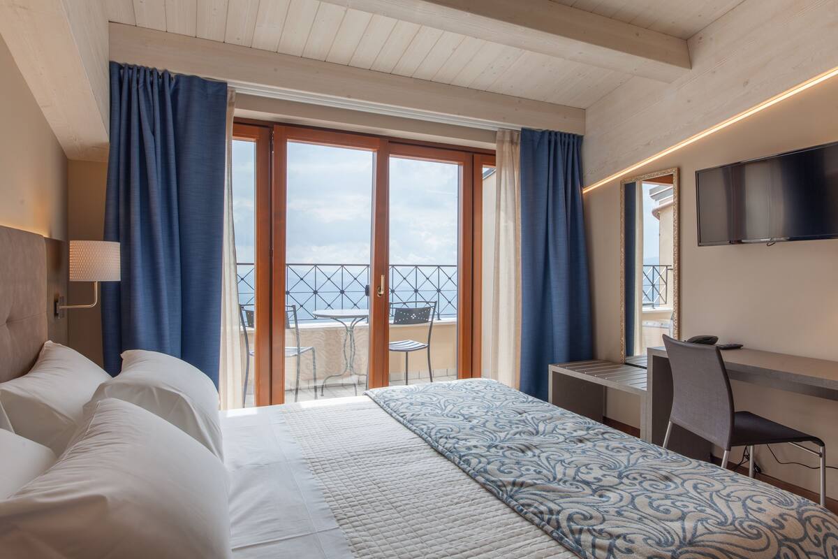 A hotel room with a bed, a desk, a chair, a TV, and a balcony with a view of the sea.