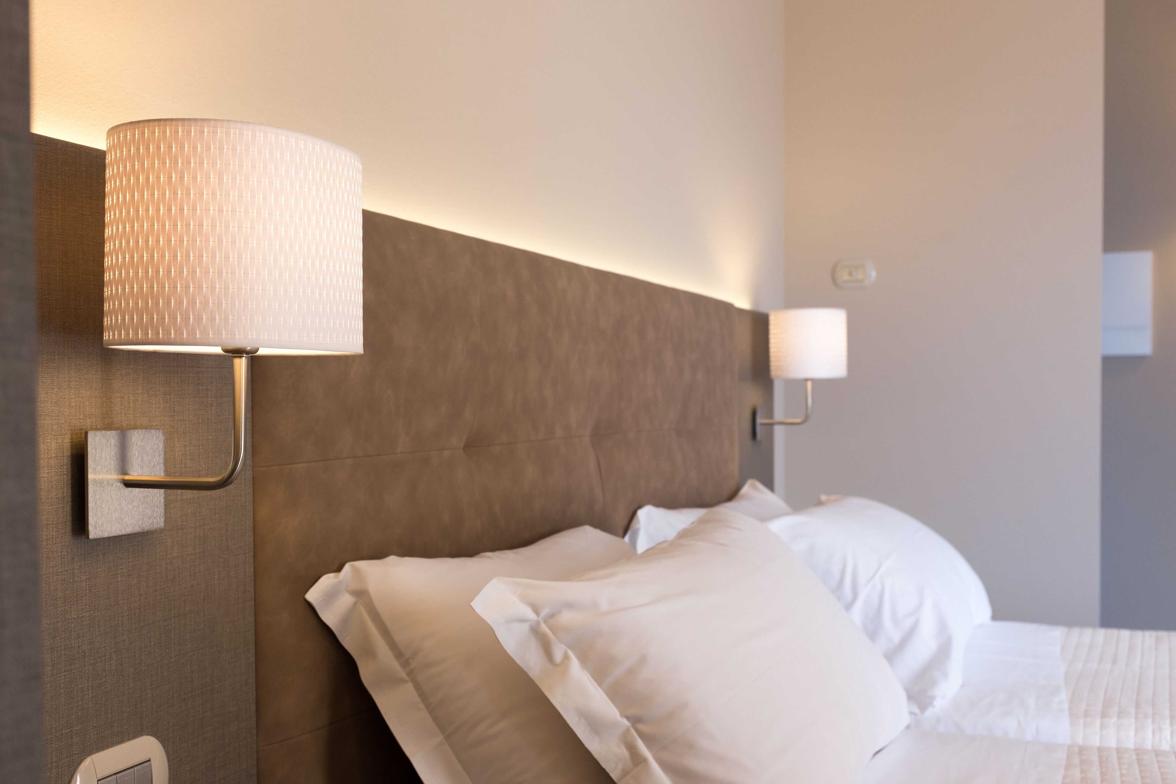 Quarto de hotel com uma cama moderna, uma luminária fixada na parede e uma mesa de cabeceira com abajur.