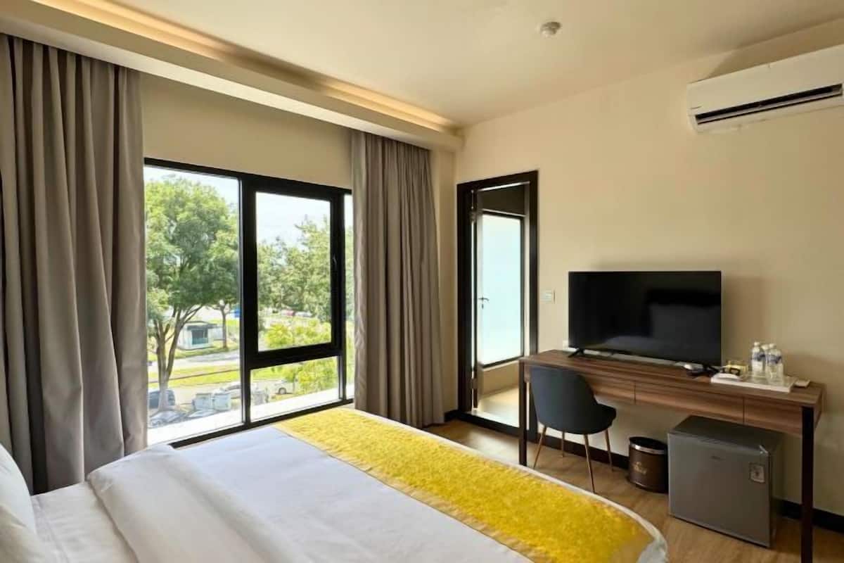 A hotel room with a bed, a desk with a TV, a small fridge, and a view of greenery outside.
