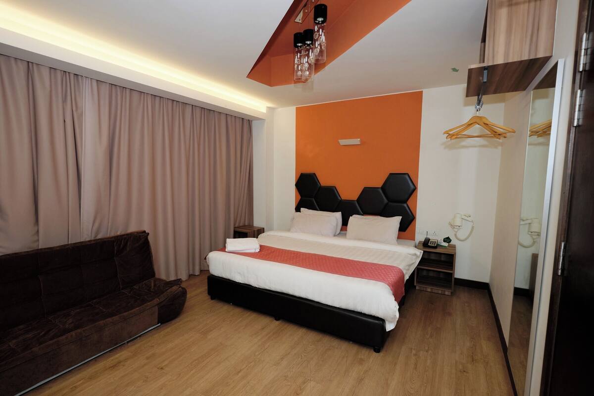 A modern hotel room with a large bed, a dark brown sofa, a wooden floor, and a mirror on one wall.