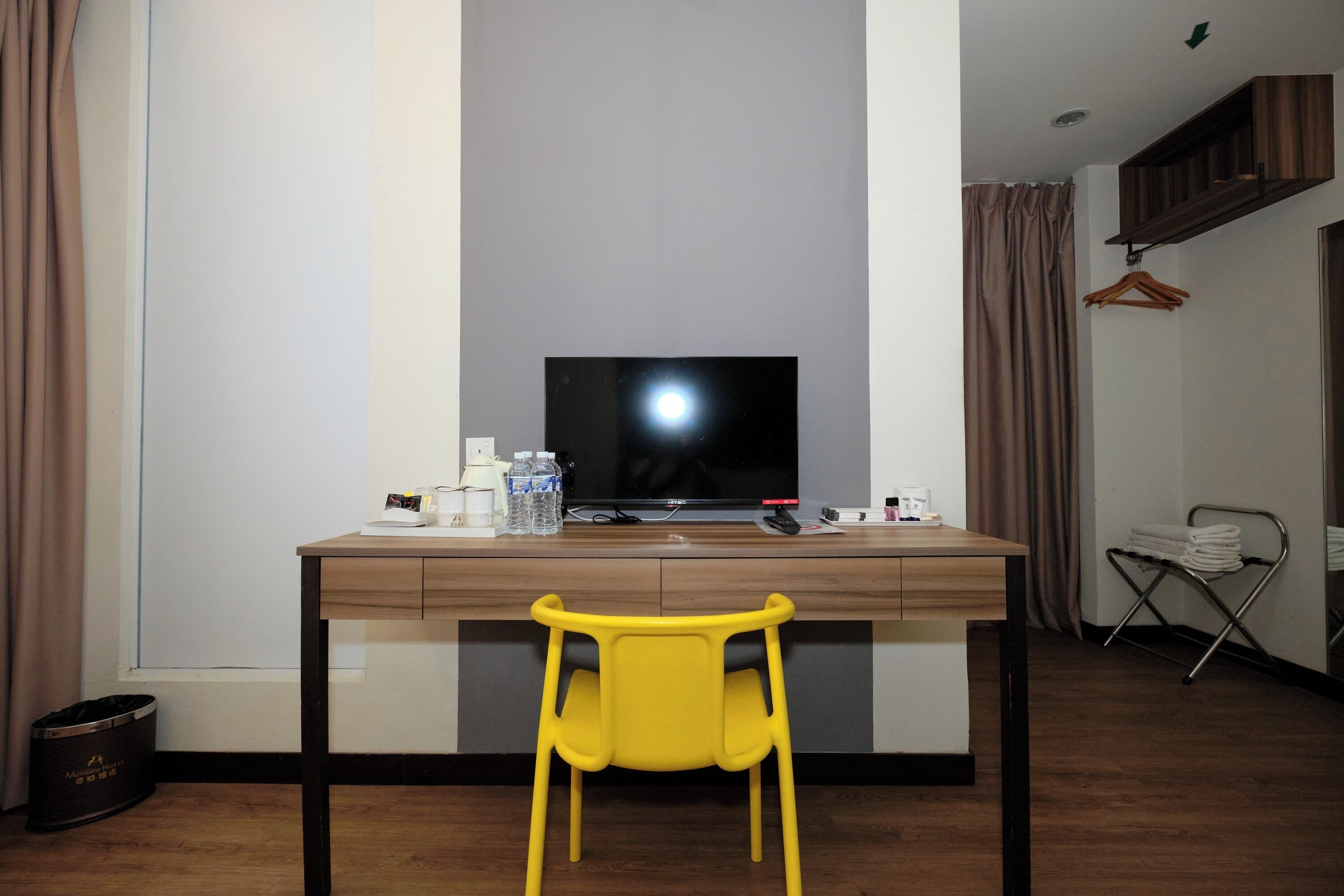 A room with a wooden desk, a yellow chair, a television, a small table with bottles and a remote control, and a hanging clothes rack.