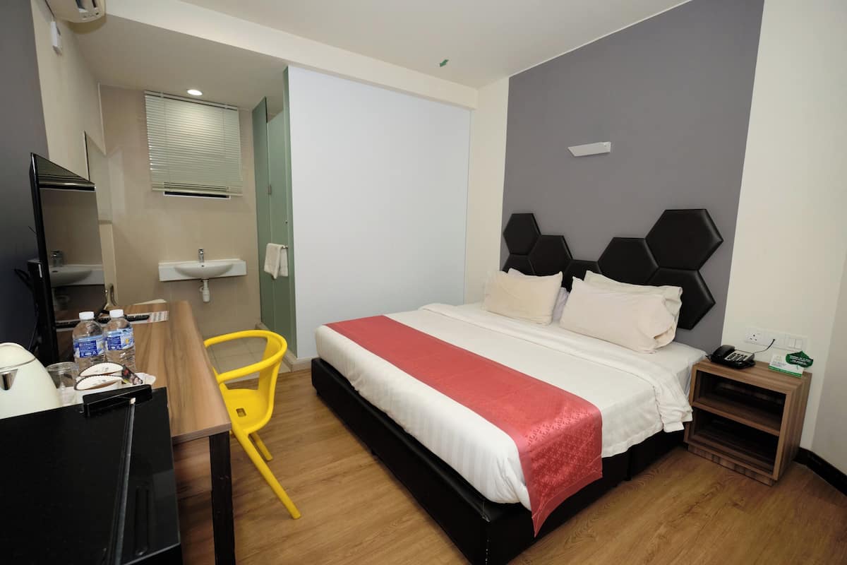 A hotel room with a bed, bedside table, chair, and a small desk with a refrigerator.
