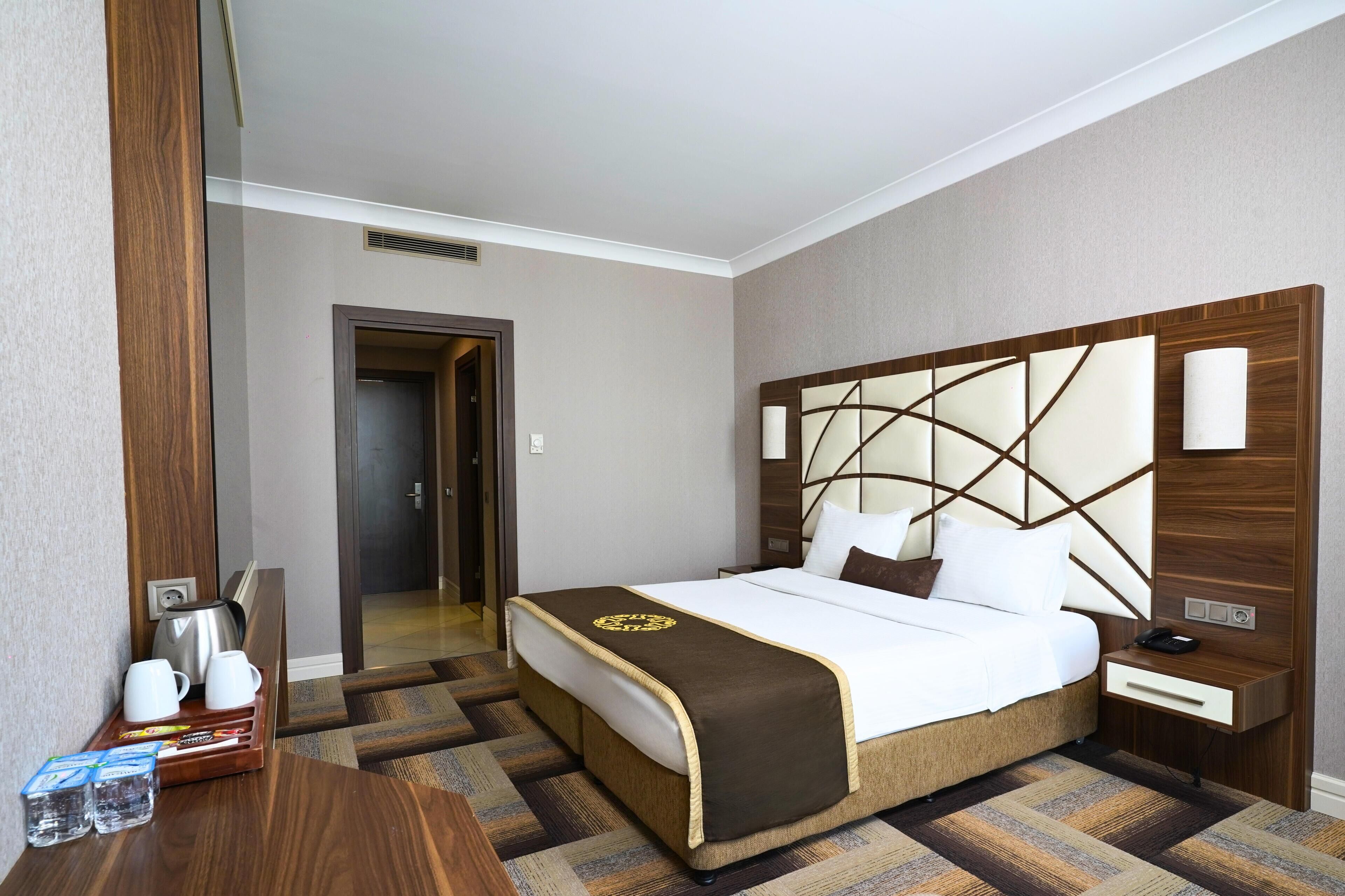 A hotel room with a large bed, a wooden headboard, a nightstand, a desk with a kettle and cups, and a tray with water bottles and snacks.