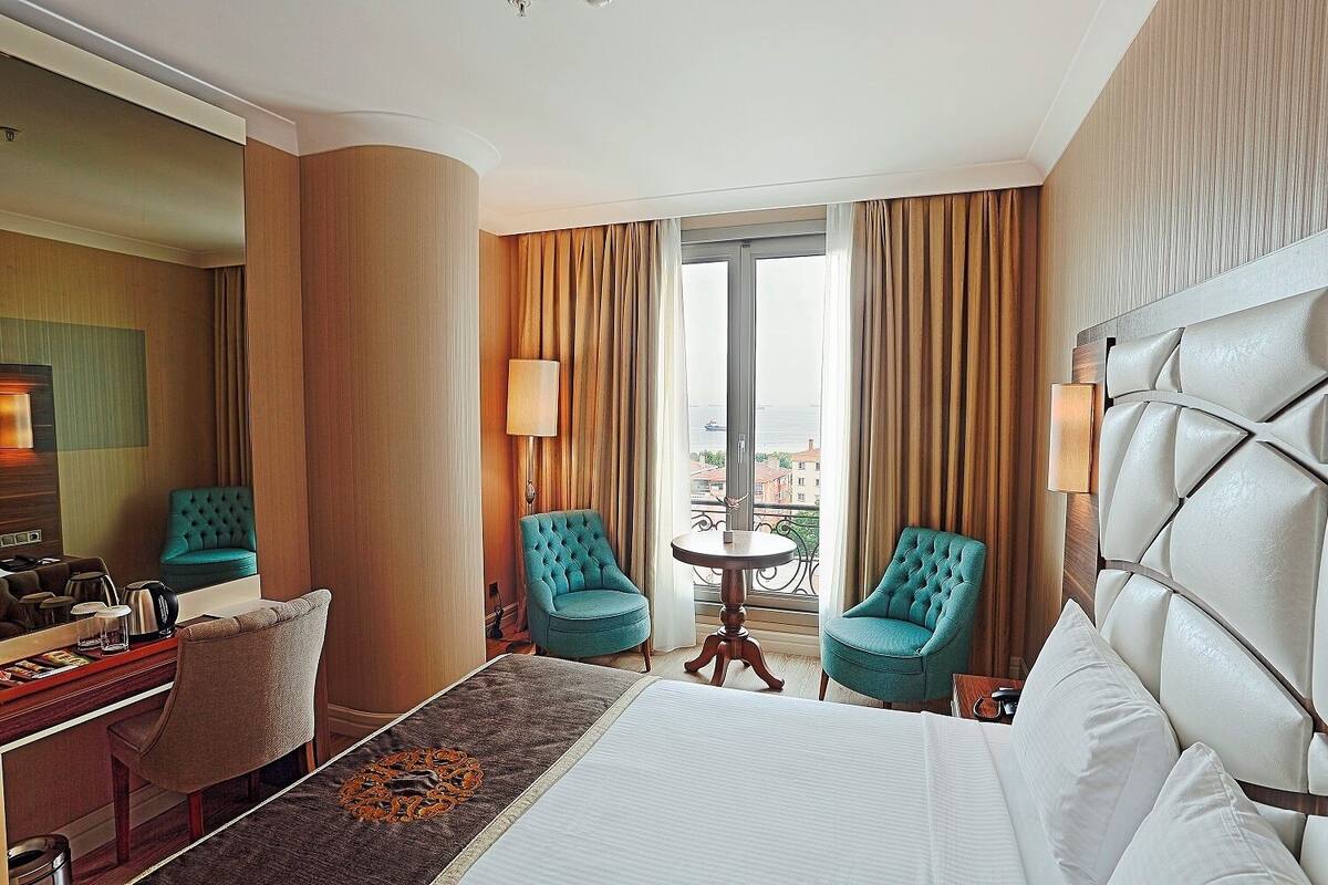 A hotel room with a large bed, two armchairs, a small table, a desk with a chair, a large mirror, and a view of the city through the window.
