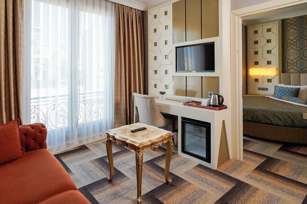 A hotel room with a sofa, a small table with a remote control, a TV mounted on a wall, a bed, and a minibar.