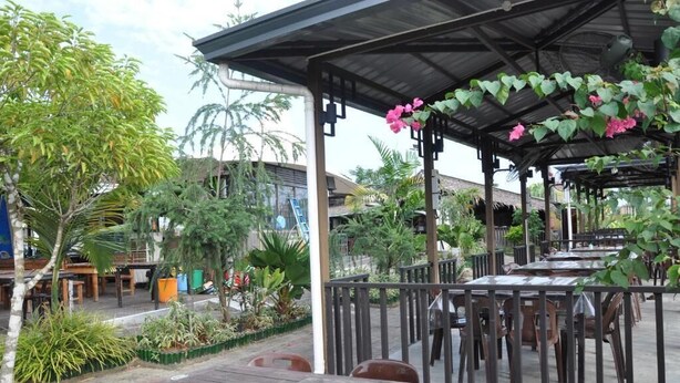 Ein Restaurantbereich im Freien mit Tischen und Stühlen, umgeben von viel Grün und einem überdachten Sitzbereich.