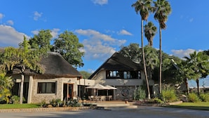 A two-story house with a thatched roof and a stone facade, surrounded by lush greenery and palm trees. - La Rochelle (Tsumeb)