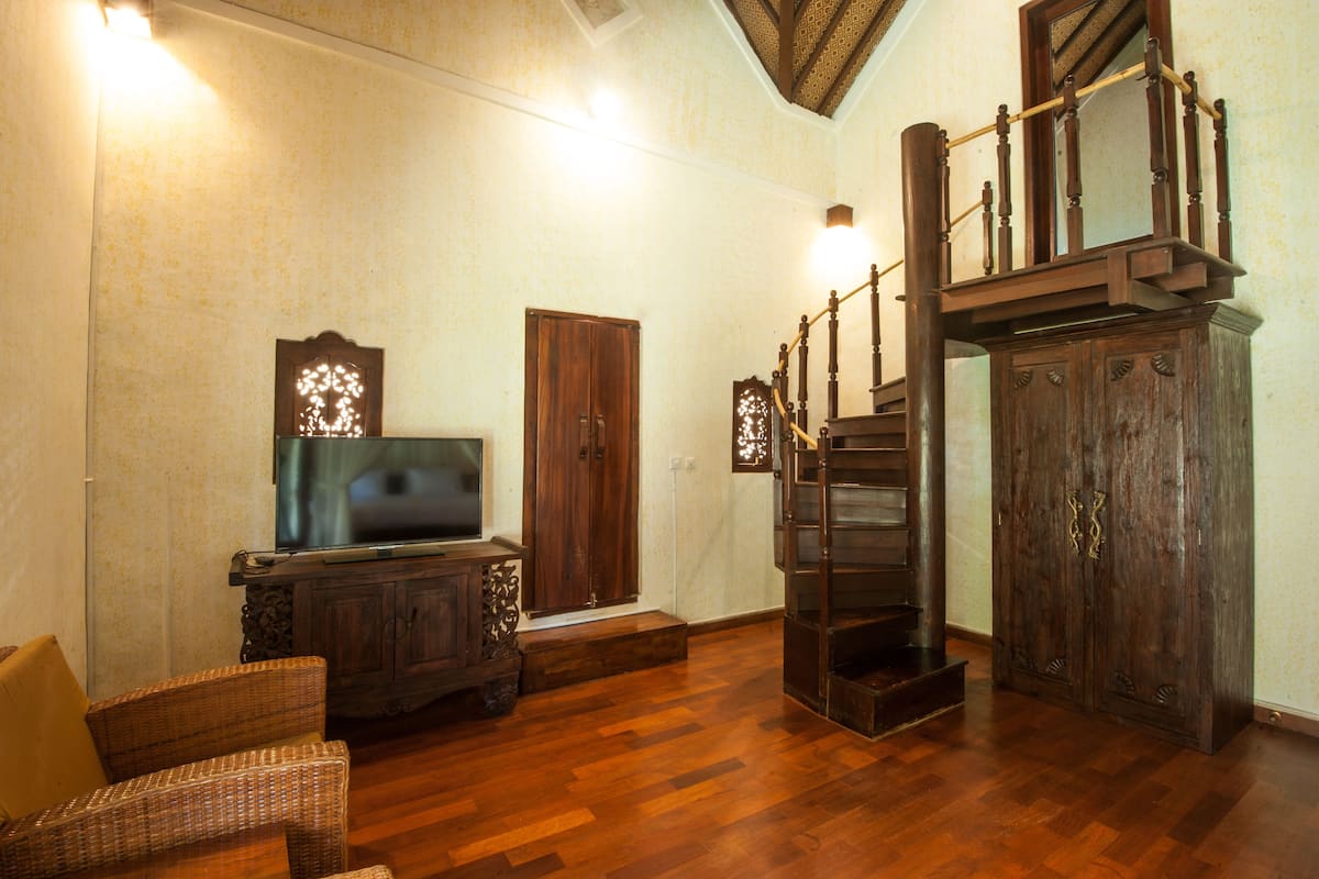 Un salón amplio con una escalera de madera, un televisor sobre un mueble bajo y una butaca de mimbre.
