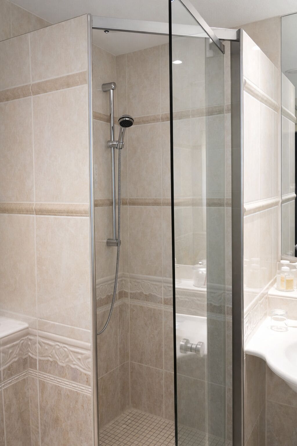Ducha con mampara de vidrio, cabezal de ducha manual, paredes de azulejos color beige y encimera blanca con artículos de aseo.