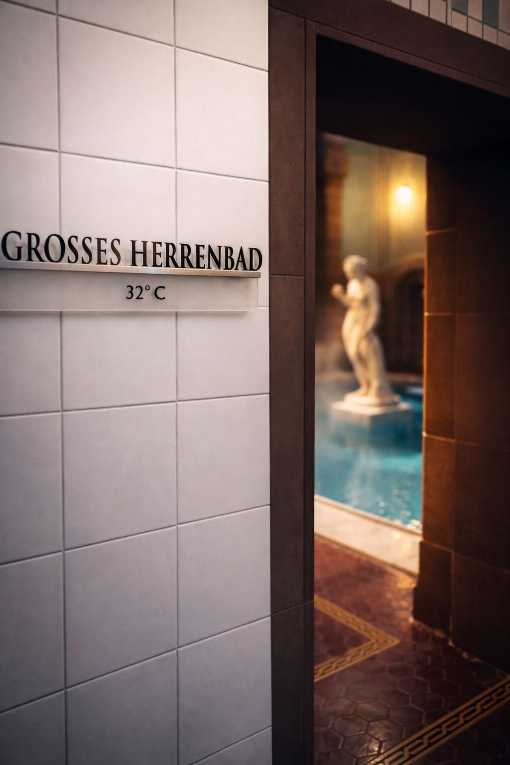 A sign reading "GROSSES HERRENBAD 32°C" with a view of a swimming pool and a statue in the background.