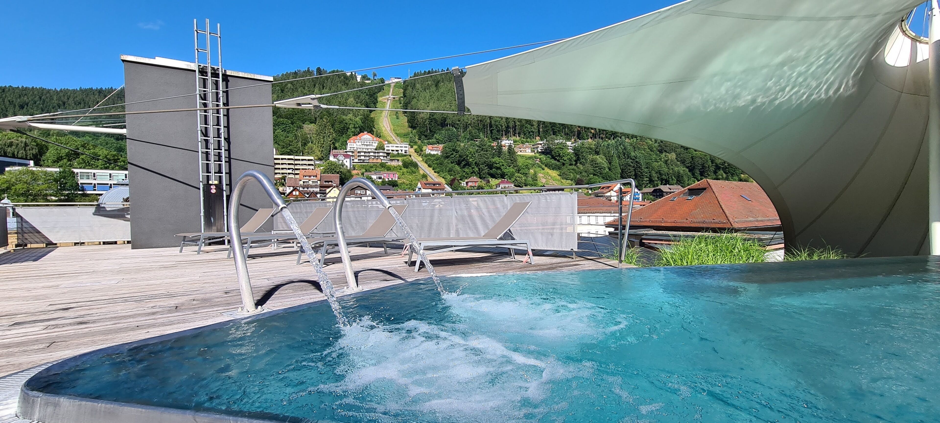Une piscine dotée d’une terrasse en bois, d’une échelle et offrant une vue sur une colline parsemée de bâtiments.