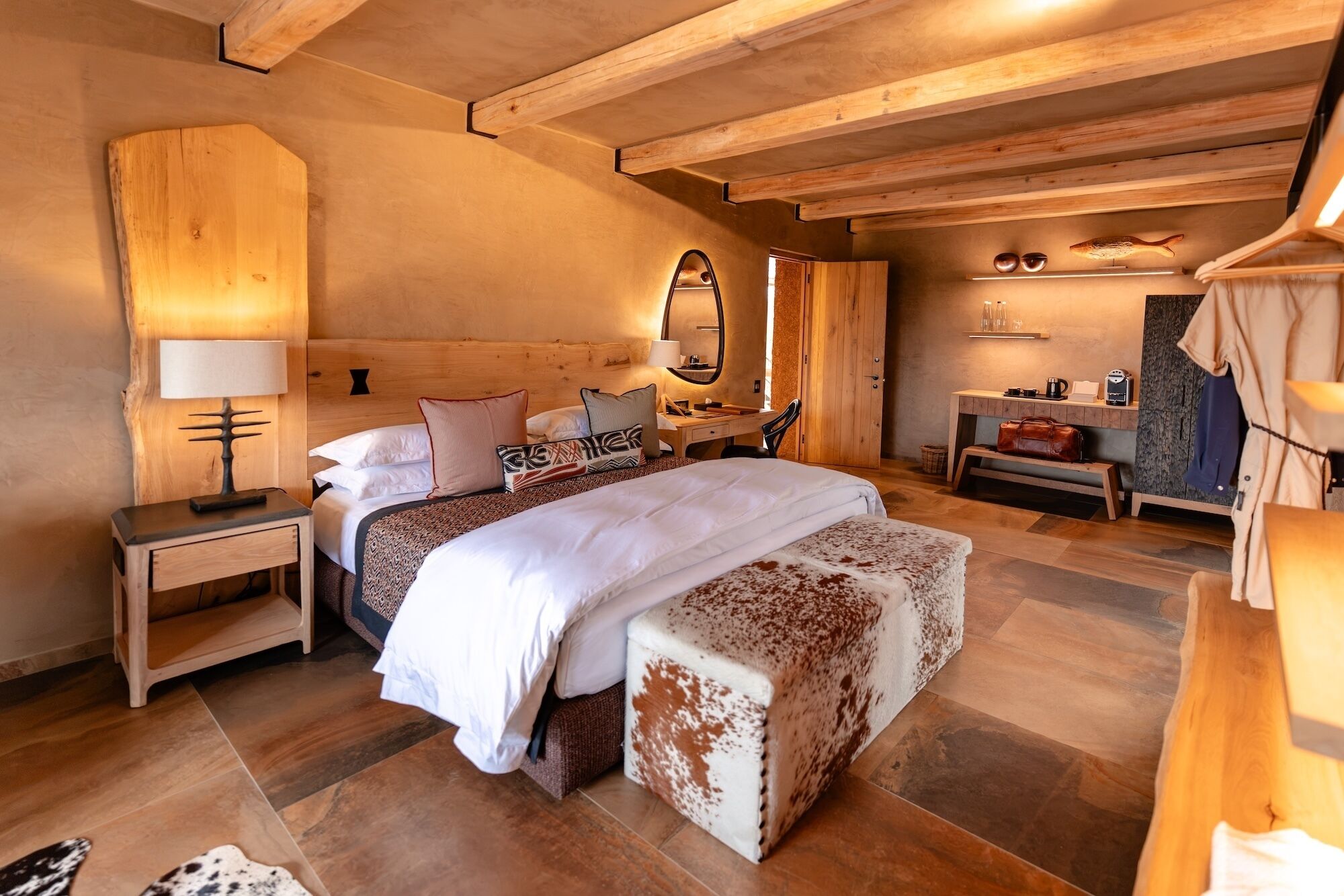 A cozy bedroom with a wooden ceiling, a large bed with multiple pillows, a bedside table, a bench, and a small desk with a chair.
