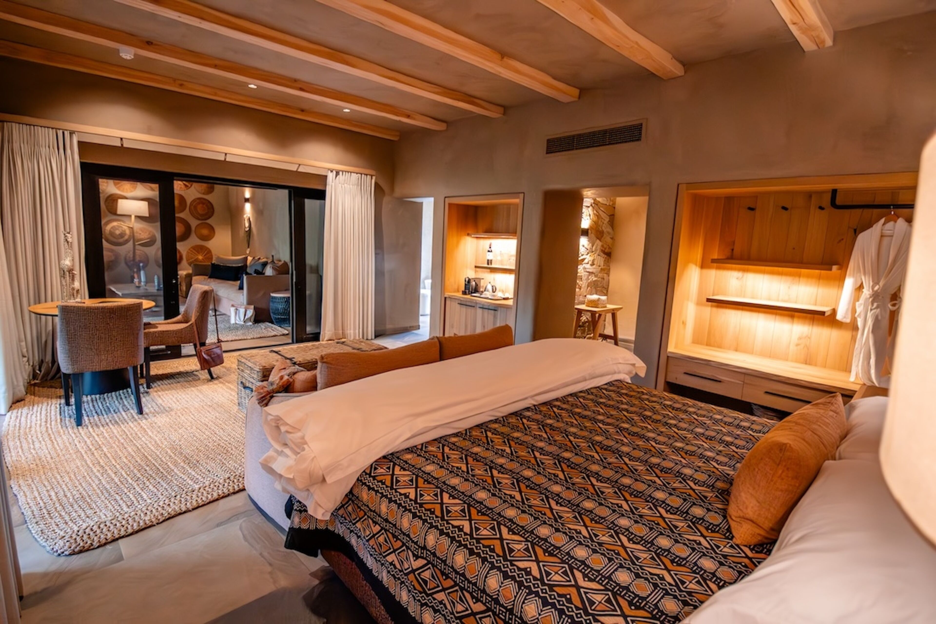A modern hotel room with a large bed, a sitting area with a chair and a small table, a bathroom with a wooden vanity, and a large window.