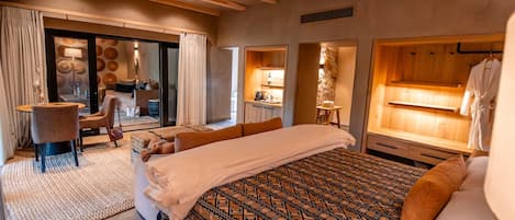 A modern hotel room with a large bed, a sitting area with a chair and a small table, a bathroom with a wooden vanity, and a large window.