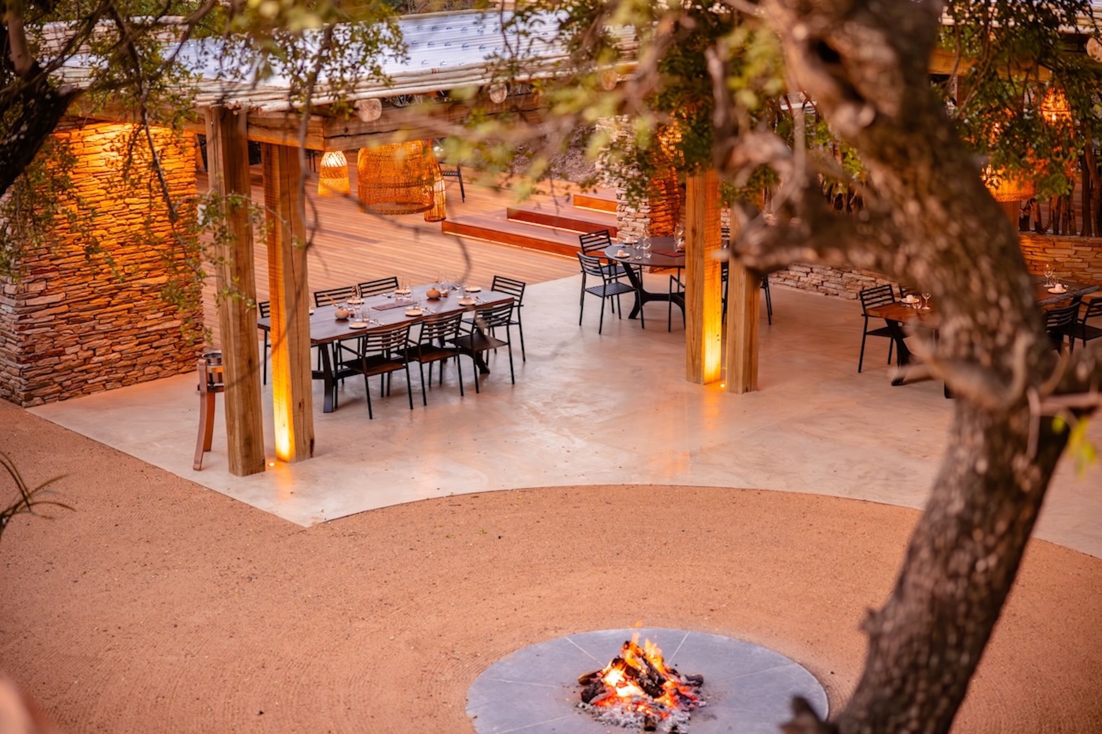 Un área de comedor al aire libre con paredes de piedra, mesas y sillas de madera, y un brasero en el centro.
