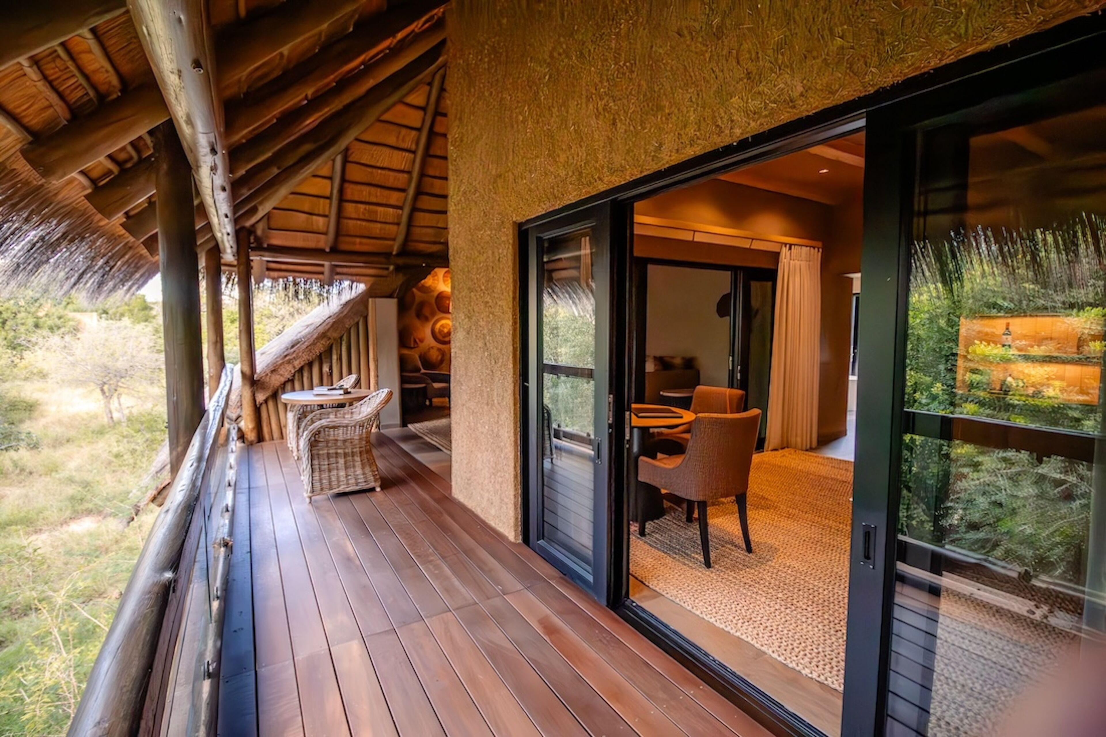 A wooden deck with a thatched roof, a dining area with chairs and a table, and a glass door leading to a room with a view of greenery.