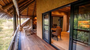 A wooden deck with a thatched roof, a dining area with chairs and a table, and a glass door leading to a room with a view of greenery.