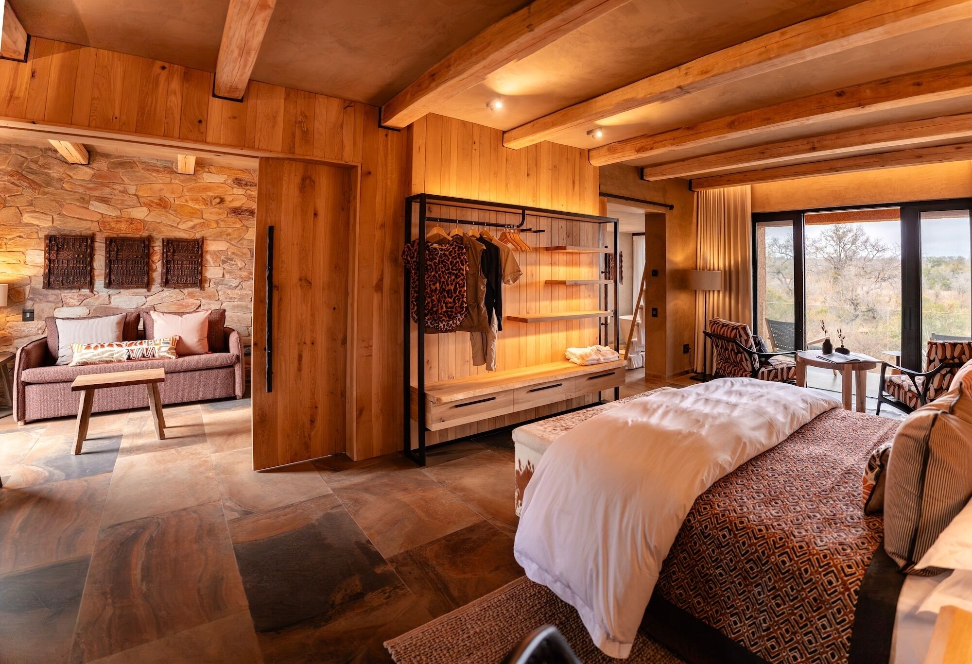 A spacious room with a large bed, a seating area with a sofa and armchairs, a wooden wardrobe, and a stone wall.