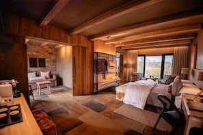A spacious room with a large bed, a seating area with a sofa and chairs, a wooden wardrobe, and a stone wall. - Kapama River Lodge (Hoedspruit)