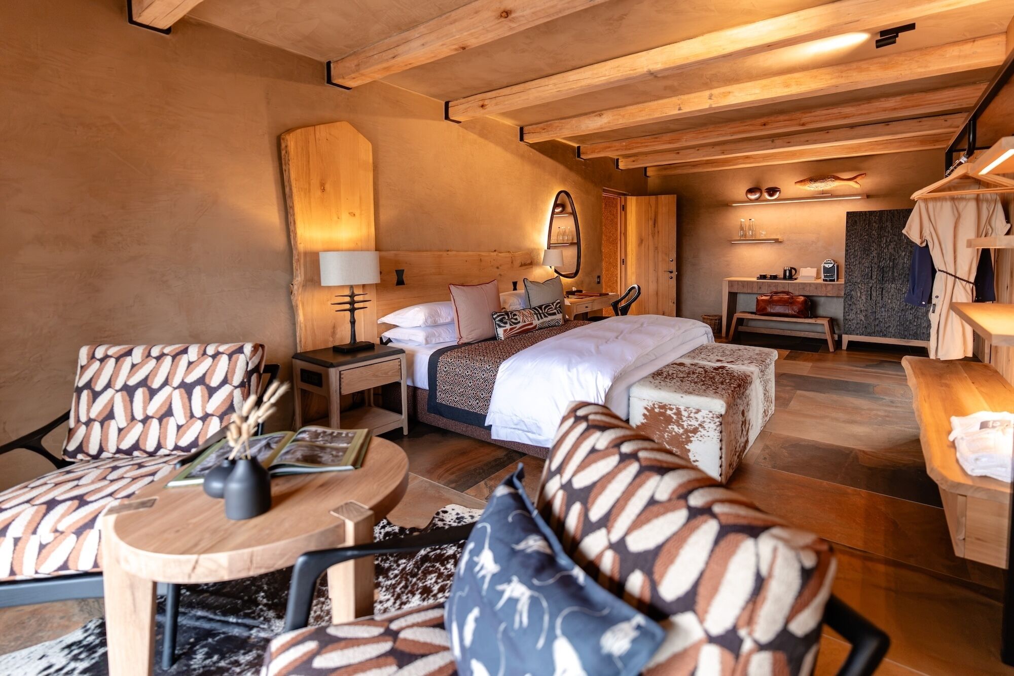Ein gemütliches Schlafzimmer mit einem großen Bett, einem Holz-Kopfteil, einem Nachttisch mit Lampe, einem kleinen Schreibtisch und einem Sessel mit gemusterten Kissen.