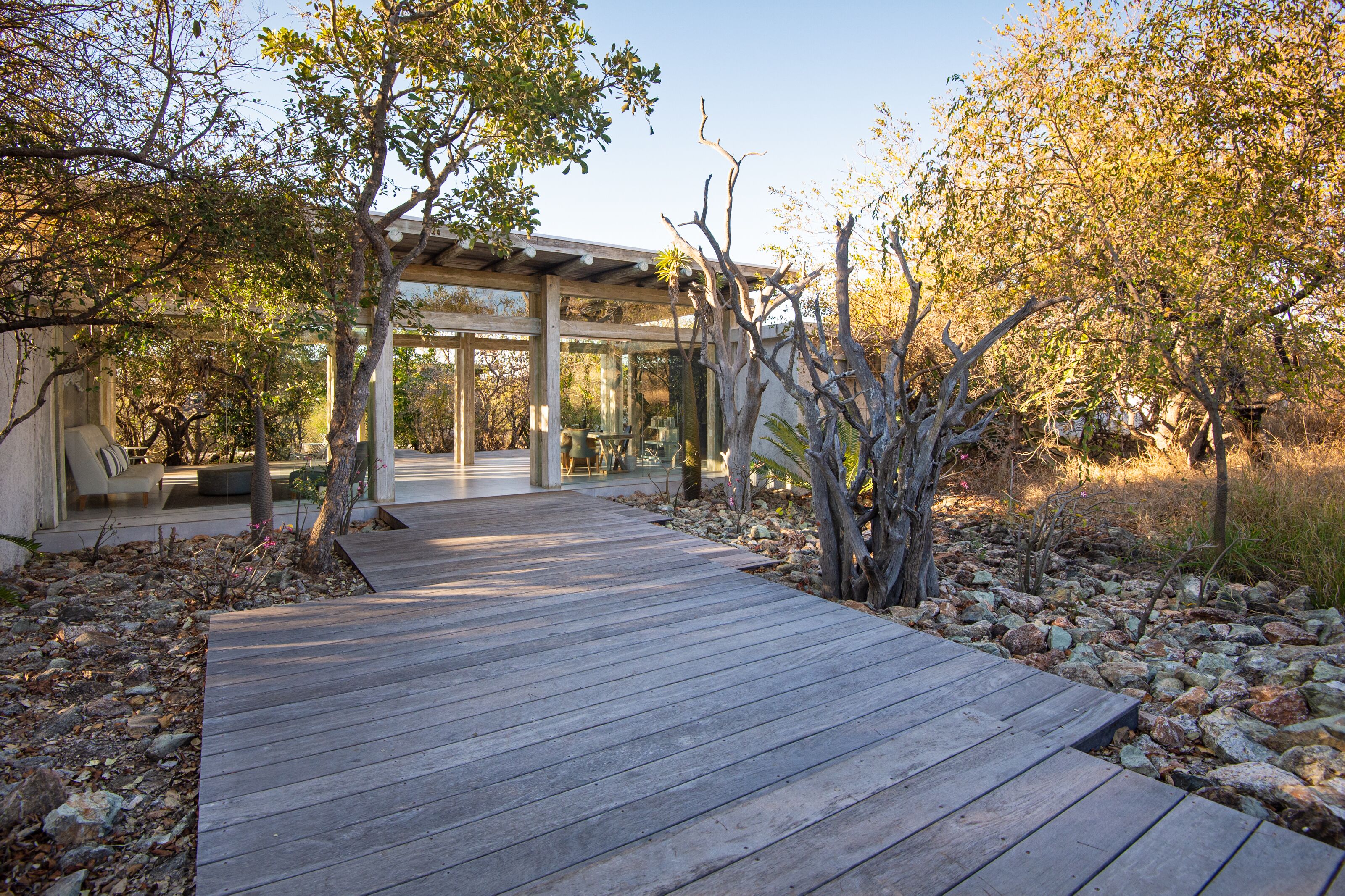 A wooden deck leading to a covered outdoor seating area with a glass enclosure, surrounded by trees and rocks.