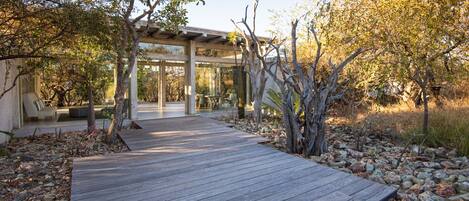 A wooden deck leading to a covered outdoor seating area with a glass enclosure, surrounded by trees and rocks.