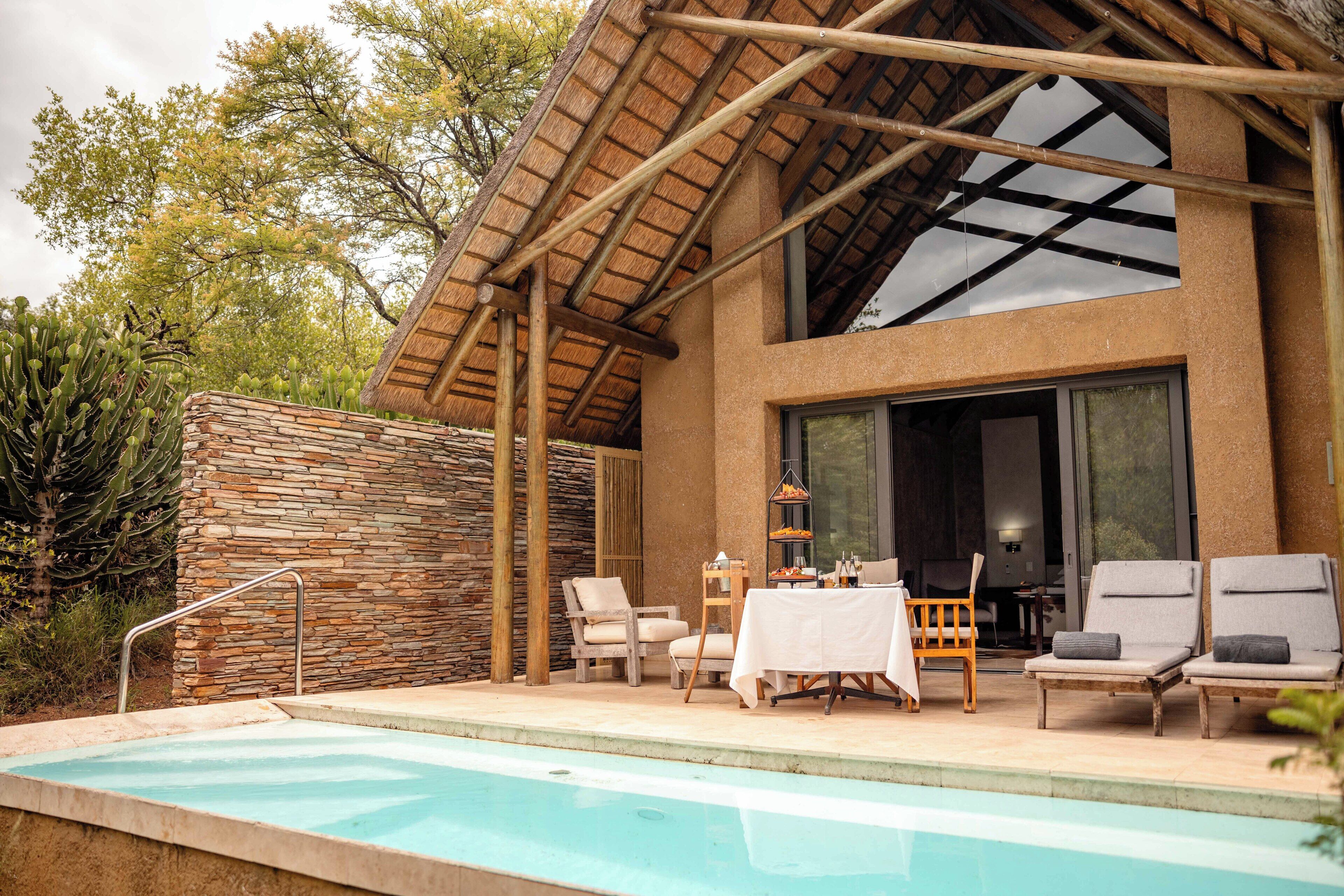 A poolside area with a stone wall, lounge chairs, a table with a white tablecloth, and a thatched roof structure.