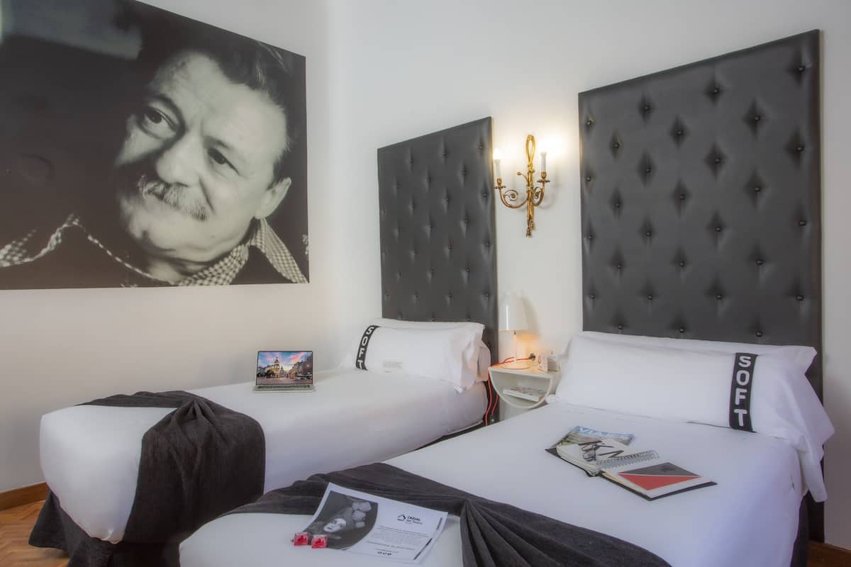 Una habitación de hotel con dos camas, un retrato grande en la pared, una mesita de noche con una lámpara, y un aplique de pared.