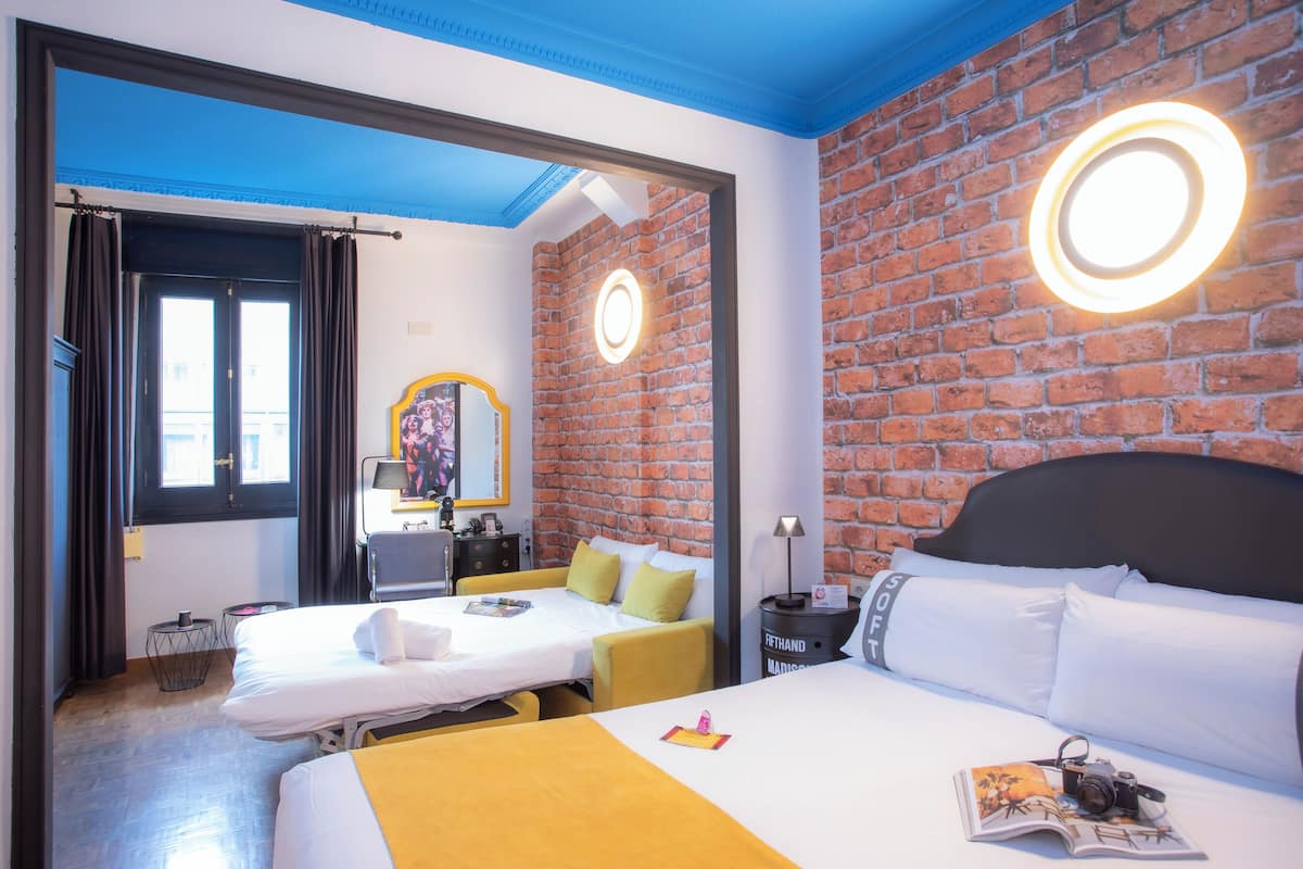 Habitación de hotel con pared de ladrillo, cama con ropa de cama blanca, un sillón amarillo y una mesita de noche con lámpara.