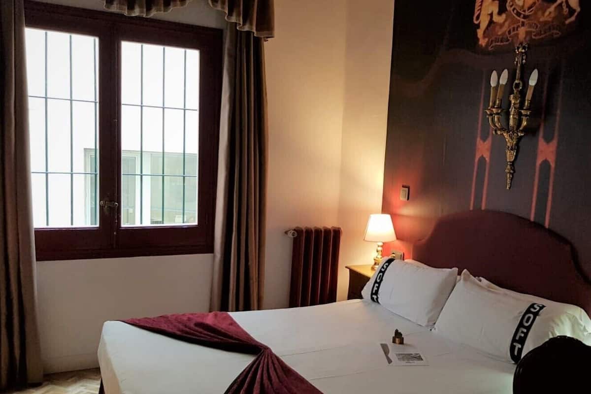 Habitación de hotel con una cama grande, una ventana con cortinas, una mesita de noche con lámpara y una pared con un escudo de armas.