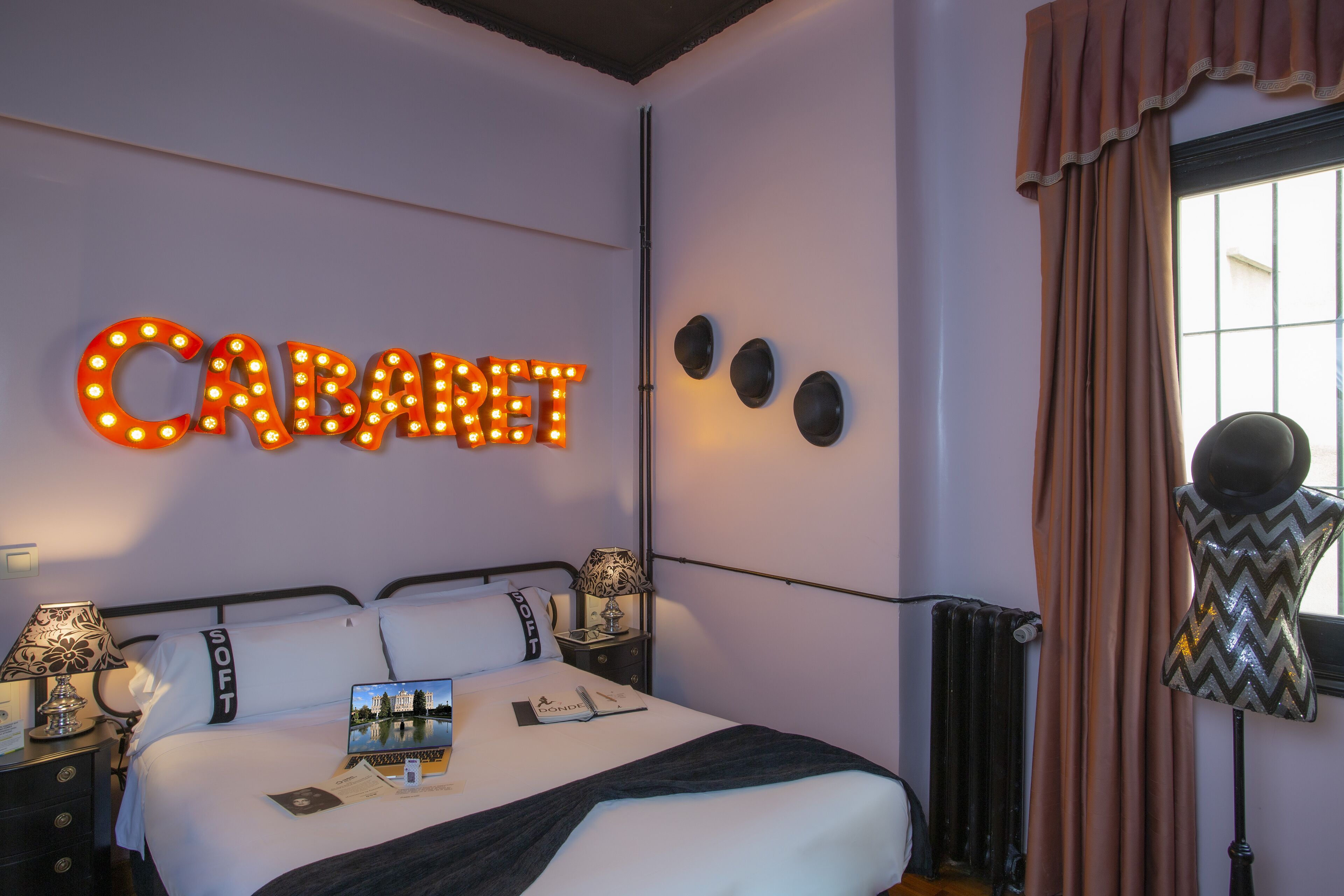 Une chambre d’hôtel avec un lit, une table de chevet, une lampe, un miroir et une fenêtre avec des rideaux.