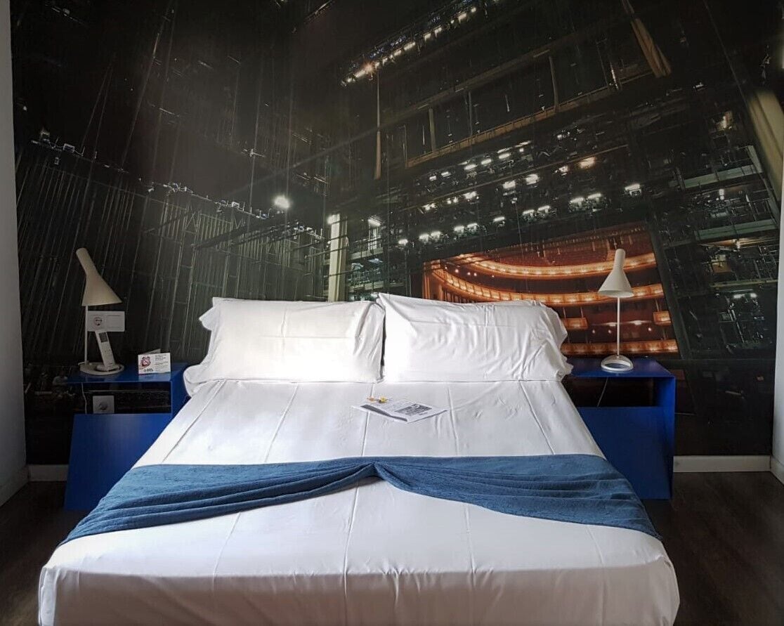A bed with white linens, a blue throw blanket, and a book on the bed. There are two bedside tables with lamps. The room has a large window with a view of an indoor space with lights.