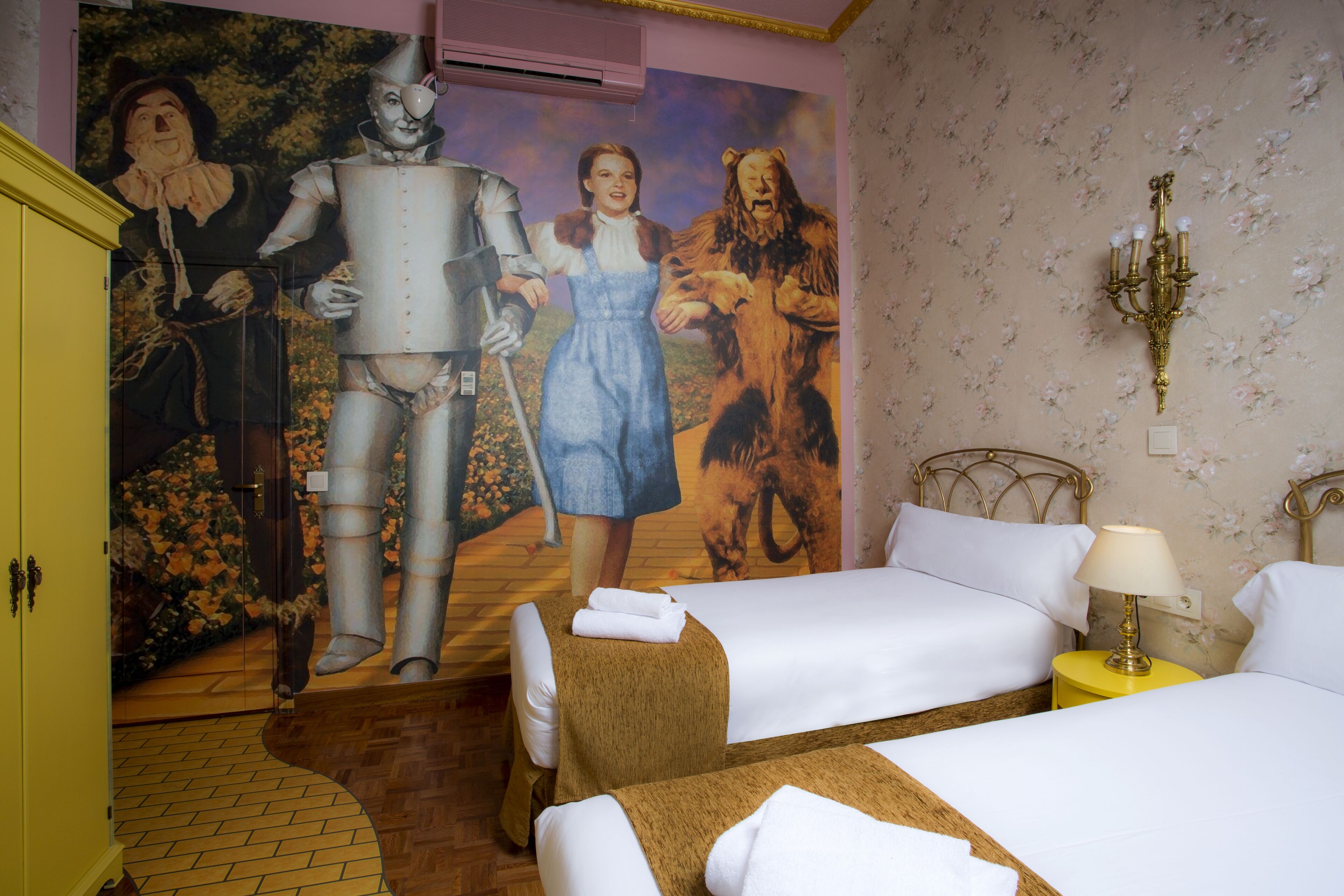 A hotel room with two beds, a mural on the wall, a chandelier, and a yellow cabinet.