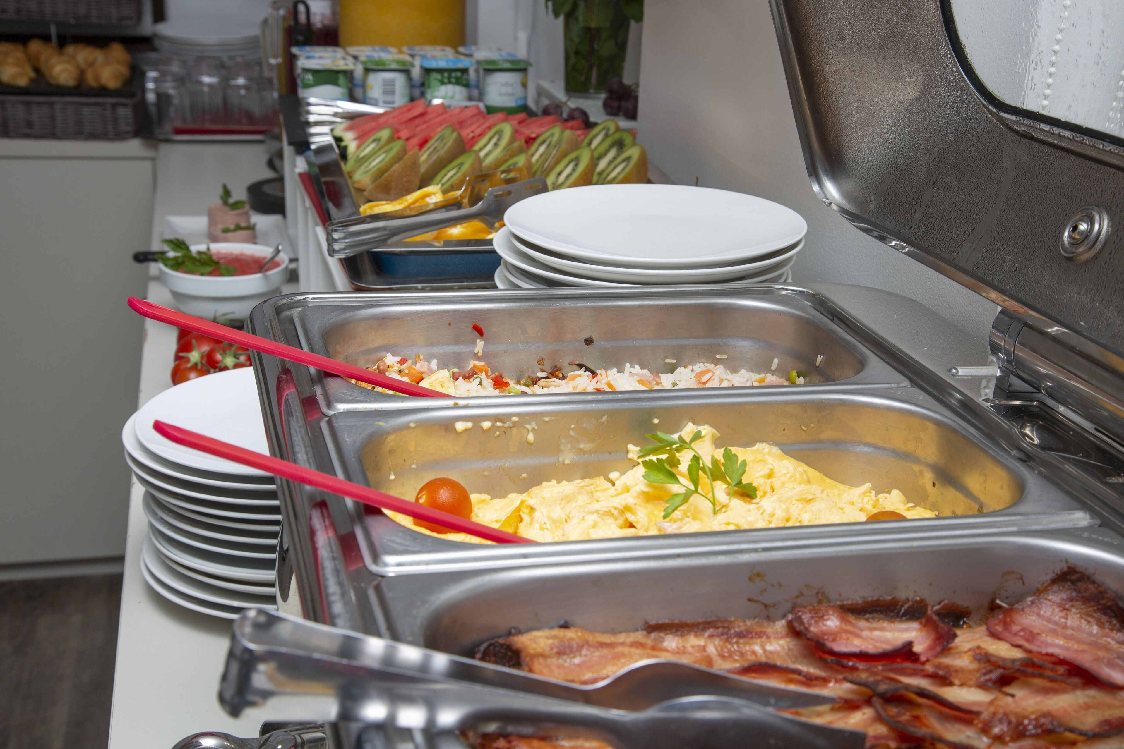Un bufé con diferentes opciones de comida, incluyendo tocino, huevos revueltos y frutas frescas.