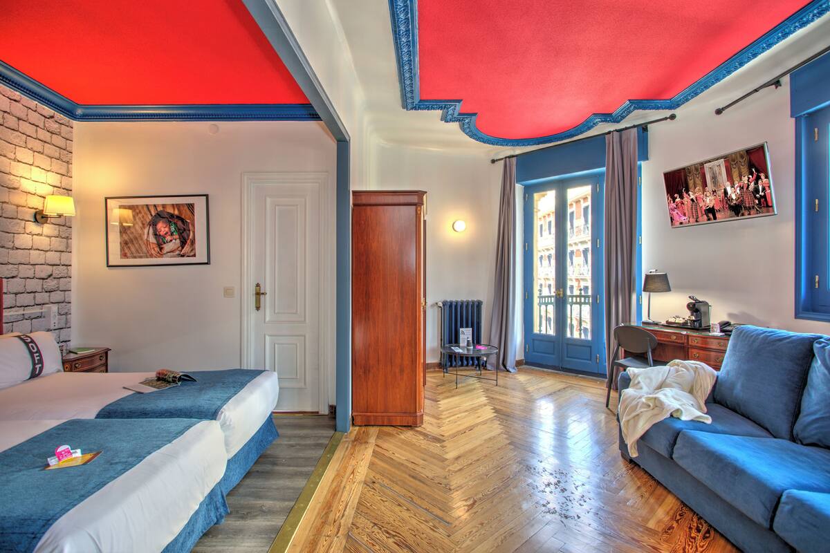 Una habitación de hotel con dos camas, un sofá azul, suelo de madera y techo rojo.