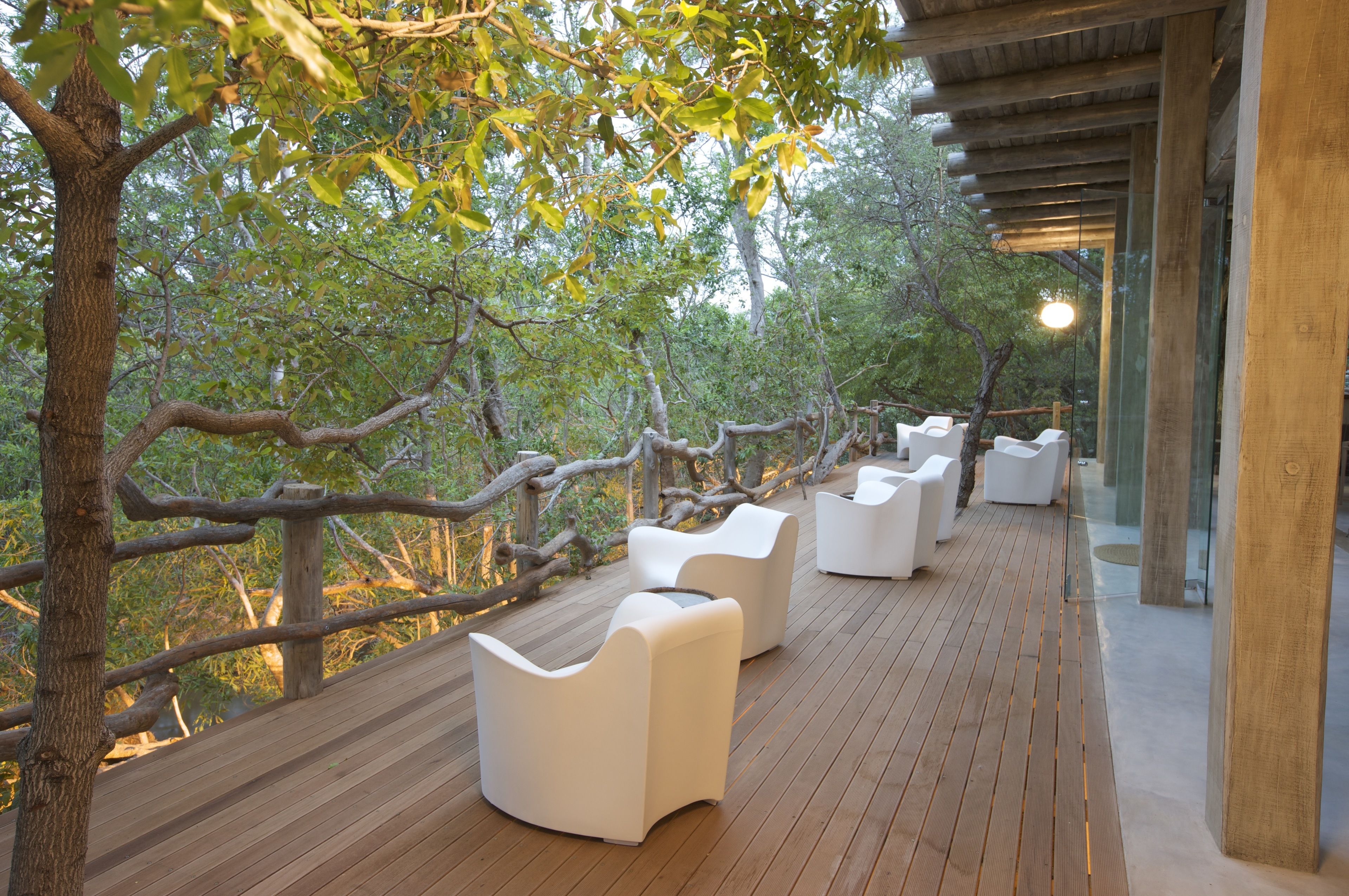 Une terrasse en bois avec des meubles d’extérieur blancs et des murs en verre donnant sur une zone boisée.