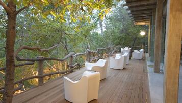 Une terrasse en bois avec des meubles d’extérieur blancs et des murs en verre donnant sur une zone boisée.