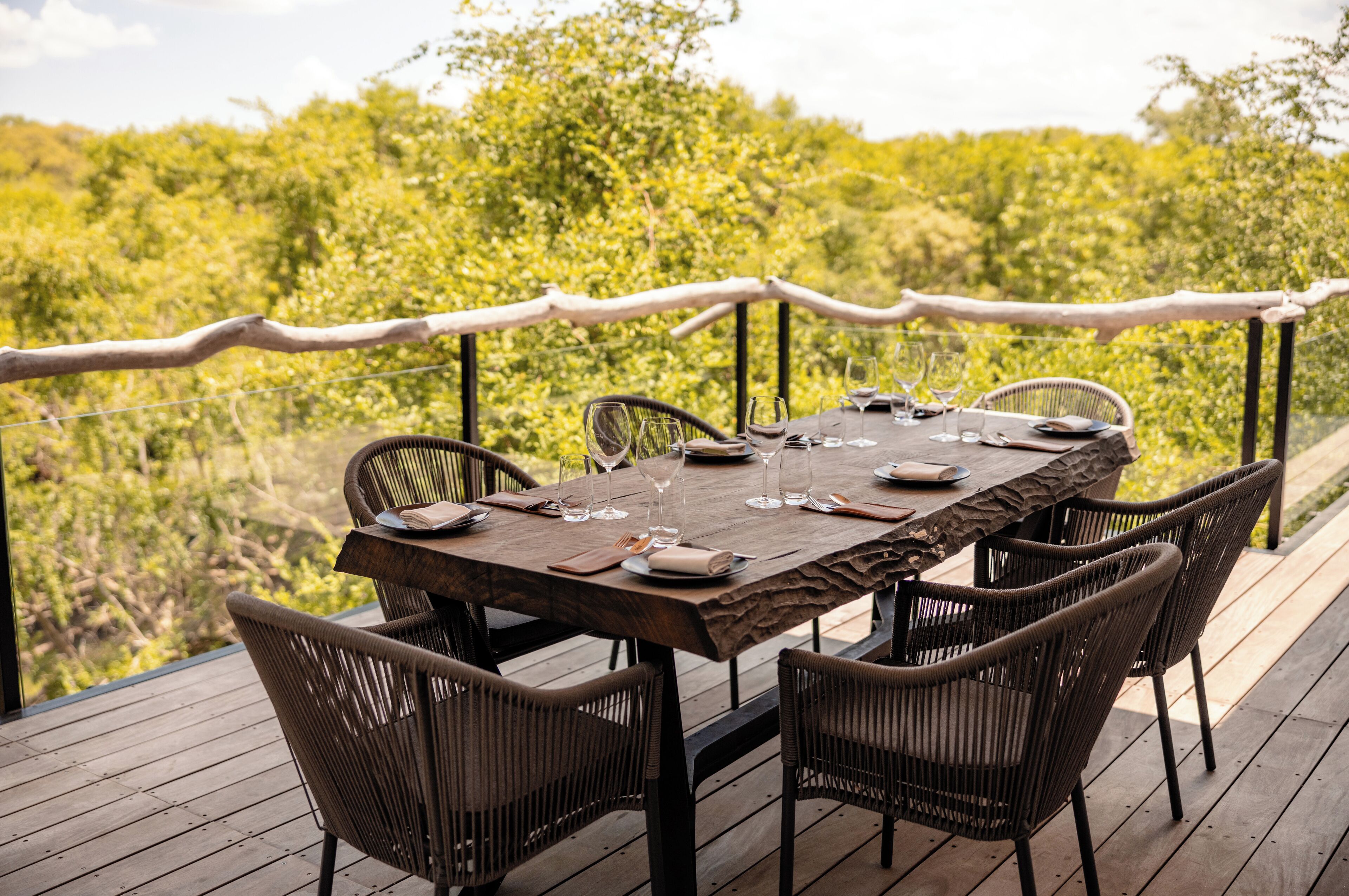 Un área de comedor al aire libre con una mesa de madera y sillas de mimbre, rodeada por una terraza de madera y vistas a un follaje exuberante.