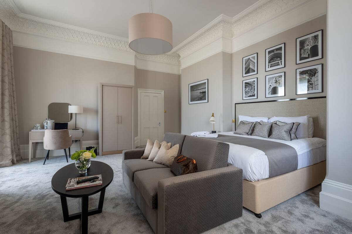 A bedroom with a large bed, a sofa, a small table with a vase and a book, and a desk with a lamp.