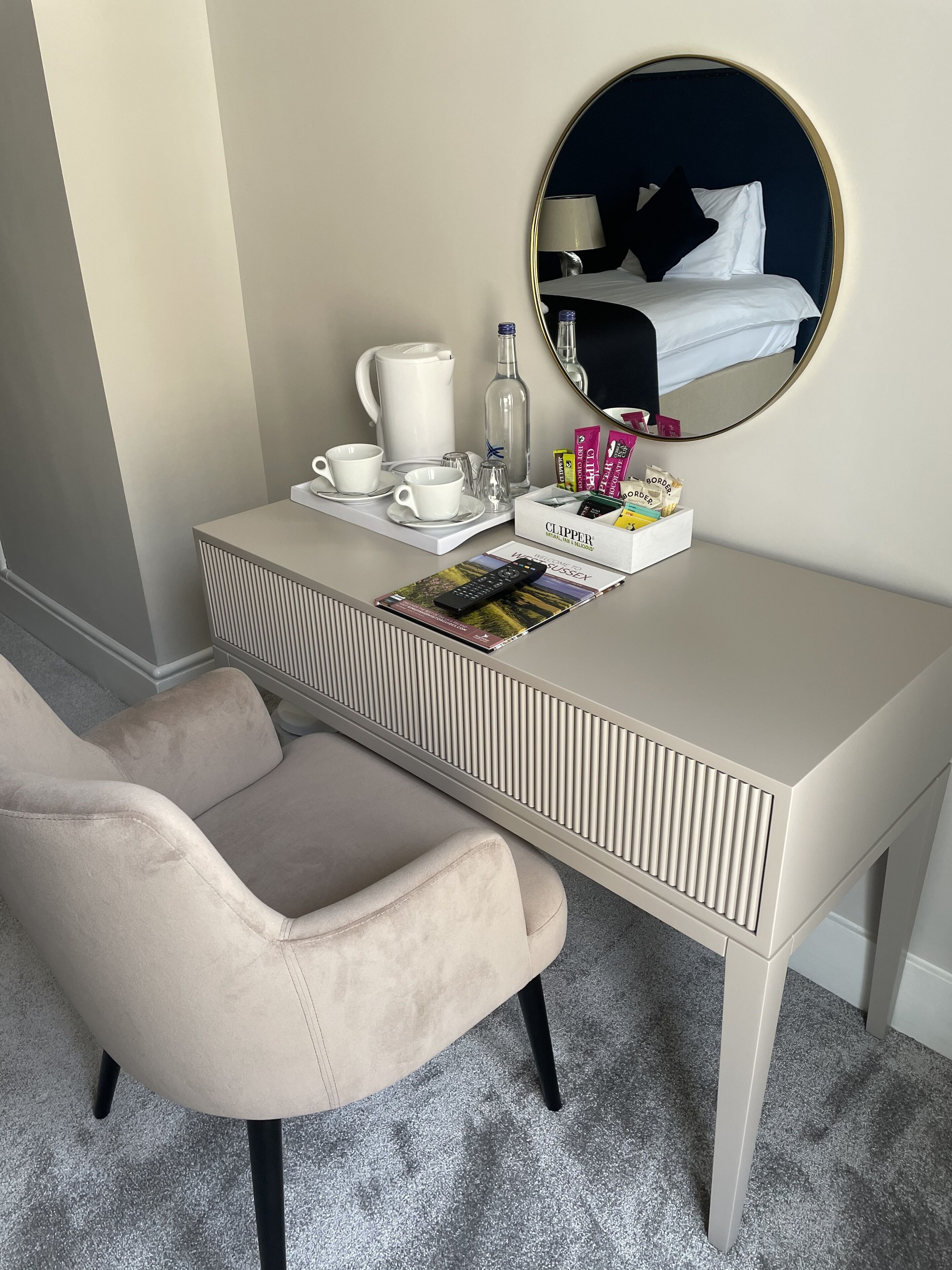 Une chambre d’hôtel moderne avec un bureau blanc, un fauteuil beige, un miroir rond et une table de chevet équipée d’une lampe, d’une machine à café et d’articles de toilette.