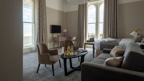 A hotel room with a bed, a sofa, a dining table with a bottle and glasses, a TV, and large windows with curtains. - The Burlington Hotel (Worthing)